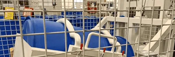 Automated drum shaker with a blue drum mounted in a steel safety cage on the production floor